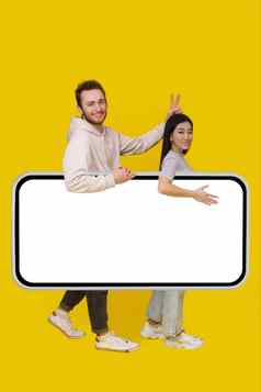 携带巨大的智能手机白色屏幕英俊的的家伙集角手势亚洲女孩移动应用程序广告兴奋微笑相机孤立的黄色的背景产品放置