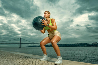 女人健身穿锻炼重医学球在户外运动女孩适合身体运动服装摆姿势天空背景