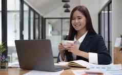 微笑年轻的亚洲女商人持有咖啡杯子移动PC办公室