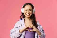 特写镜头肖像迷人的可爱的亚洲女孩听音乐耳机显示心手势胸部爱最喜欢的艺术家歌手执行太棒了歌曲粉红色的背景