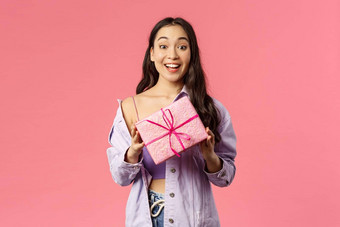 现在兴奋快乐年轻的亚洲女孩持有包装盒子礼物祝贺假期生日邀请生日聚会，派对站逗乐粉红色的背景