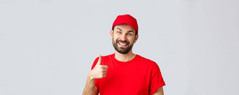 在线购物交付检疫外卖概念微笑英俊的有胡子的交付的家伙快递红色的统一的帽显示支持翘拇指保证安全订单转移