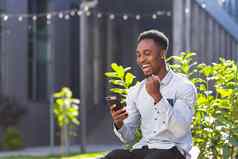 快乐非洲美国男人。休闲衣服坐着板凳上在户外移动电话