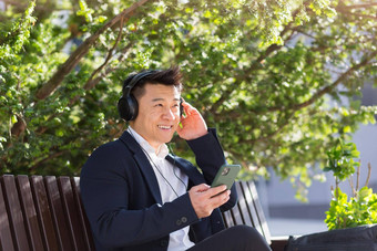 快乐的亚洲业务男人。坐着板凳上城市公园市中心听音乐