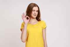 肖像满意年轻的女人棕色（的）波浪头发休闲黄色的t恤站相机显示标志手势眨眼