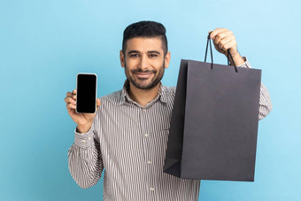 商人胡子持有白色显示智能手机黑色的纸袋在线购物