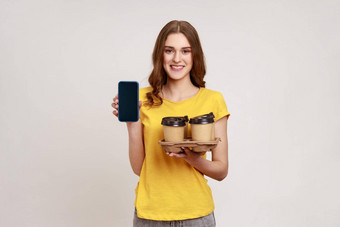 积极的微笑少年女孩棕色（的）头发黄色的t恤持有空显示智能手机持有人咖啡纸杯在线<strong>订单</strong>