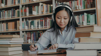 漂亮的积极的亚洲十几岁的学生耳机头听音乐坐着表格大图书馆持有书写作总结