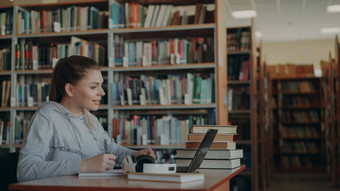 年轻的美丽的微笑学生女孩坐着桌子上大宽敞的图书馆桩书写作<strong>字帖</strong>移动PC前面