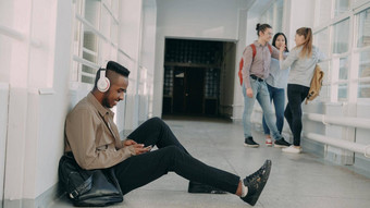 年轻的英俊的非洲美国学生坐着地板上白<strong>色</strong>走廊耳机头听音乐持有智能手机发短信<strong>同学</strong>们