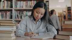 年轻的美丽的亚洲女学生坐着表格包围桩书大图书馆重写文本教科书准备好了考试