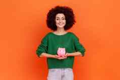 愉快的女人非洲式发型发型穿绿色休闲风格毛衣站持有粉红色的小猪银行手储蓄钱