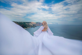 金发女郎长头发阳光明媚的海滨白色流动衣服后视图丝绸织物挥舞着风背景蓝色的天空山海滨