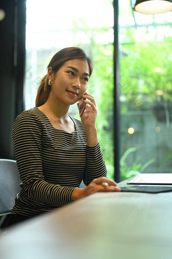 迷人的亚洲女自由职业者坐着咖啡商店会说话的移动电话