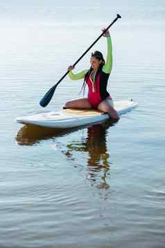 年轻的女人长发绺泳装坐着董事会女paddleboarding湖日出