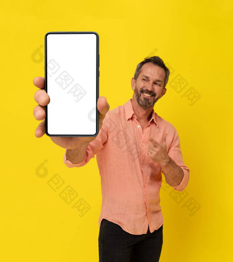 快乐中间岁的灰色头发的适合男人。持有大智能手机指出手指穿桃子衬衫孤立的黄色的成熟的肌肉男人。电话应用程序广告模拟白色屏幕