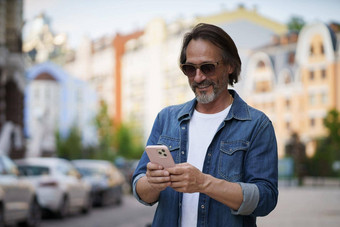 发短信发布社会媒体帖子照片成熟的英俊的男人。智能手机在户外穿牛仔布衬衫太阳镜英俊的中间岁的男人。灰色头发使照片旅行