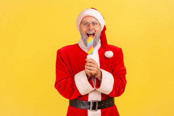 兴奋上了年纪的男人。灰色的胡子穿圣诞老人老人服装持有咬五彩缤纷的冰奶油庆祝冬天假期