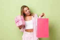 肖像时尚的金发碧眼的女孩购物假期持有礼物盒子粉红色的袋站绿色背景