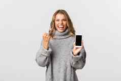 图像赢得快乐女人灰色毛衣并成就显示智能手机屏幕使拳头泵白色背景