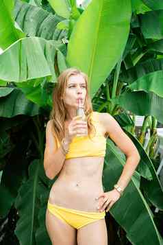 年轻的高加索人女人黄色的swuimsuit喝饮料站热带棕榈叶子