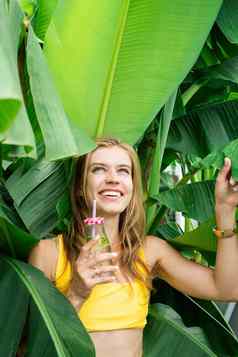 年轻的高加索人女人黄色的swuimsuit喝饮料站热带棕榈叶子