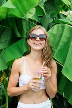 年轻的高加索人女人白色swuimsuit喝饮料站热带棕榈叶子