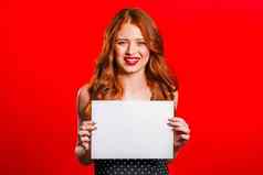 漂亮的女人持有白色纸海报复制空间微笑时尚的女孩红色的头发工作室背景