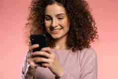女孩卷曲的头发有趣的化妆智能手机冲浪互联网粉红色的工作室背景现代技术应用程序社会网络