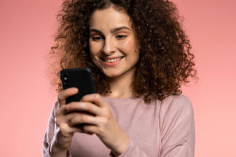 女孩卷曲的头发有趣的化妆智能手机冲浪互联网粉红色的工作室背景现代技术应用程序社会网络