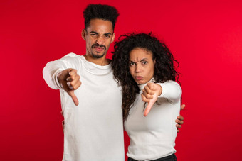 年轻的非洲夫妇站红色的工作室背景表达不满显示拇指手势相机肖像男人。女人标志不喜欢