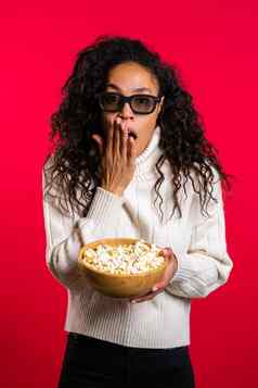 年轻的非洲女人眼镜看恐怖电影吃爆米花红色的工作室背景害怕女孩卷曲的头发出乎意外情节电影
