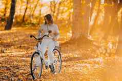 美丽的肖像年轻的女人骑自行车秋天公园阳光明媚的一天金叶子森林时尚的夫人古董白色自行车健康的生活方式审美场景