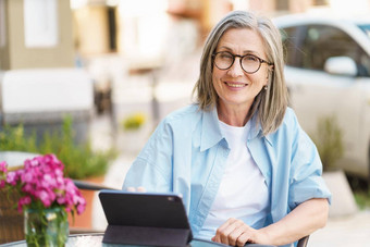 成熟的灰色头发的欧洲女人数字平板电脑坐着表格<strong>小街</strong>咖啡馆成熟的银头发女人工作在户外城市咖啡馆穿白色t恤蓝色的衬衫