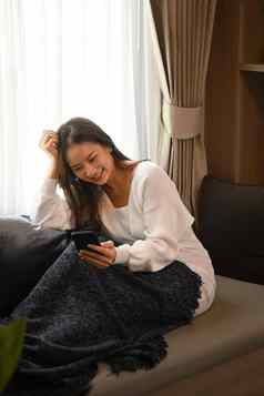 年轻的女人温暖的毛衣享受聊天在线检查社会媒体聪明的电话坐着沙发上首页