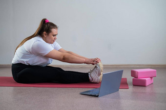 年轻的高加索人脂肪女人弯曲体育席看培训视频移动PC胖乎乎的女孩伸展运动远程视频沟通