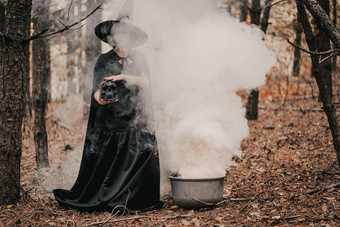 黑色的女巫持有头骨手抚摸秋天森林背景恐怖万圣节动漫真人秀假期魔法概念