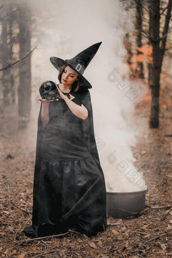 黑色的女巫持有头骨手抚摸秋天森林背景恐怖万圣节动漫真人秀假期魔法概念