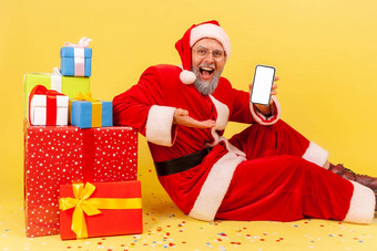 快乐上了年纪的男人。灰色的胡子穿圣诞老人老人服装展示空白白色显示细胞电话坐着一年现在盒子