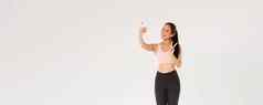完整的长度可爱的亚洲健身女孩女运动员运动服装显示和平手势采取自拍移动电话女人锻炼健身房采取图片培训会话