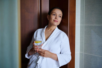 中间岁的漂亮的女人浴袍玻璃柠檬水好开始一天排毒护理健康的皮肤