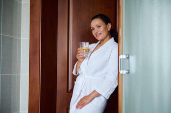 非洲美国漂亮的女人穿白色华夫格浴袍喝清洁水柠檬玻璃首页浴室