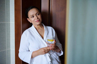 美丽的中年多民族女人白色华夫格浴袍玻璃新鲜的柠檬水排毒治疗