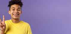特写镜头肖像英俊的乐观的年轻的十几岁的的家伙非洲式发型发型显示和平标志微笑穿黄色的t恤住乐观积极的紫色的背景