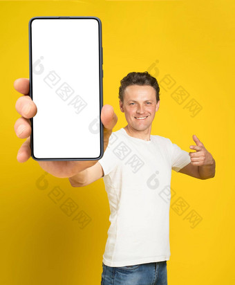持有巨大的智能手机白色屏幕英俊的男人。指出白色空屏幕穿白色t恤牛仔裤孤立的黄色的背景移动应用程序广告伟大的提供