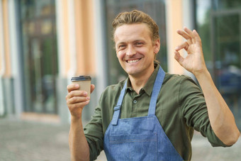 英俊的咖啡师男人。的家伙持有纸杯新鲜的使咖啡手势标志手微笑站在户外窗户商店背景自由职业者咖啡师围裙