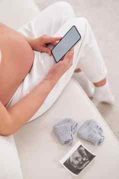 不知名的怀孕了女人坐在白色沙发持有智能手机胎儿超声波筛选