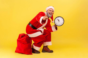 快乐积极的上了年纪的男人。灰色的胡子圣诞老人老人服装坐着红色的袋<strong>礼物</strong>持有墙时钟手指出相机