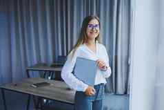 年轻的微笑业务女人站办公室窗口移动PC手女人经理眼镜白色衬衫