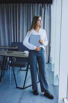 年轻的微笑业务女人站办公室窗口移动PC手女人经理眼镜白色衬衫